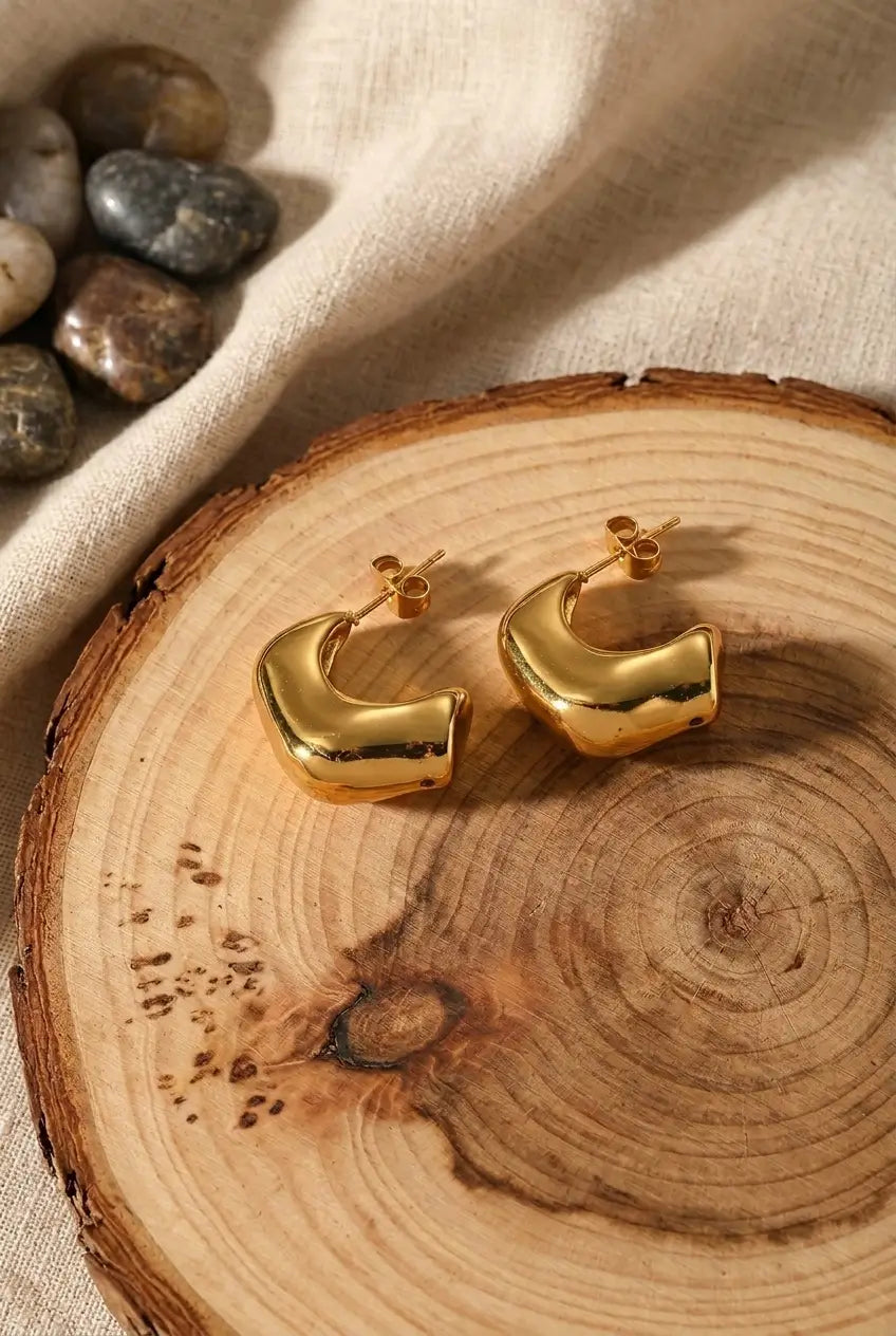 A pair of chunky gold hoop earrings with a polished, rounded modern design, displayed on a rustic wood slice with neutral fabric and stones in the background.
