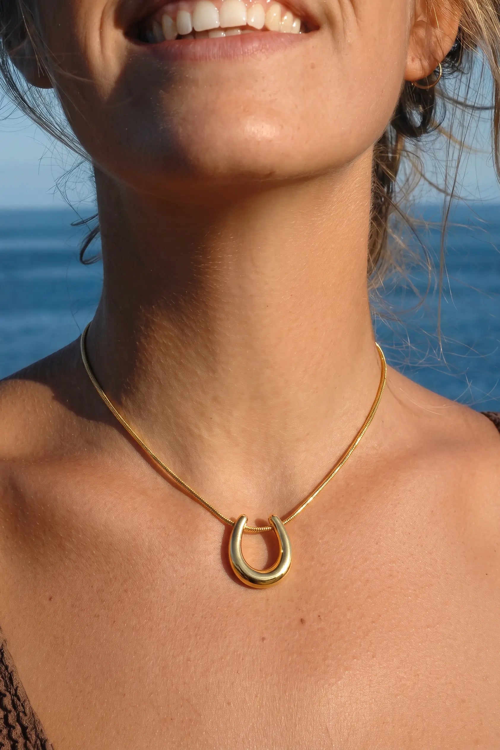 A close-up lifestyle shot of a woman wearing a gold horseshoe pendant necklace against a sunny beach background with the ocean visible.
