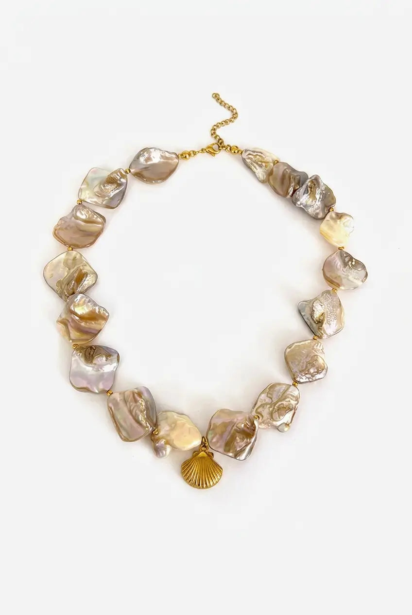 A statement collar necklace featuring irregular, organic-shaped baroque pearls in shades of beige, cream, and soft lavender, accented by a small gold shell charm and gold-tone clasp.