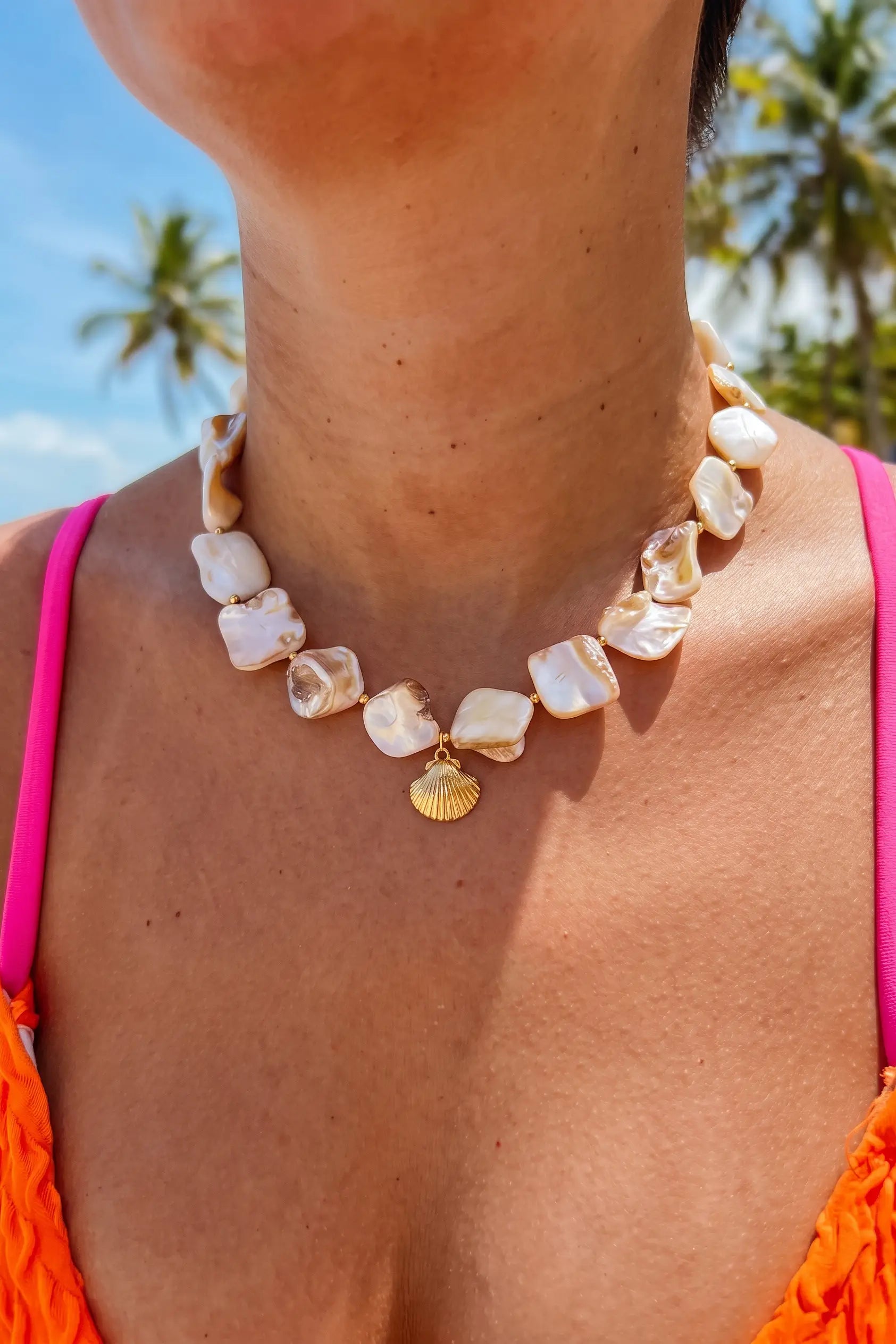 A close-up of a woman wearing a shell-bead necklace with a small gold seashell pendant, photographed outdoors with palm trees and blue sky in the background.