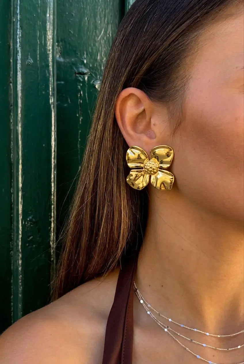 A close-up of a woman wearing a large, shiny gold flower-shaped earring, with her brown hair and shoulder visible against a dark green wooden door.