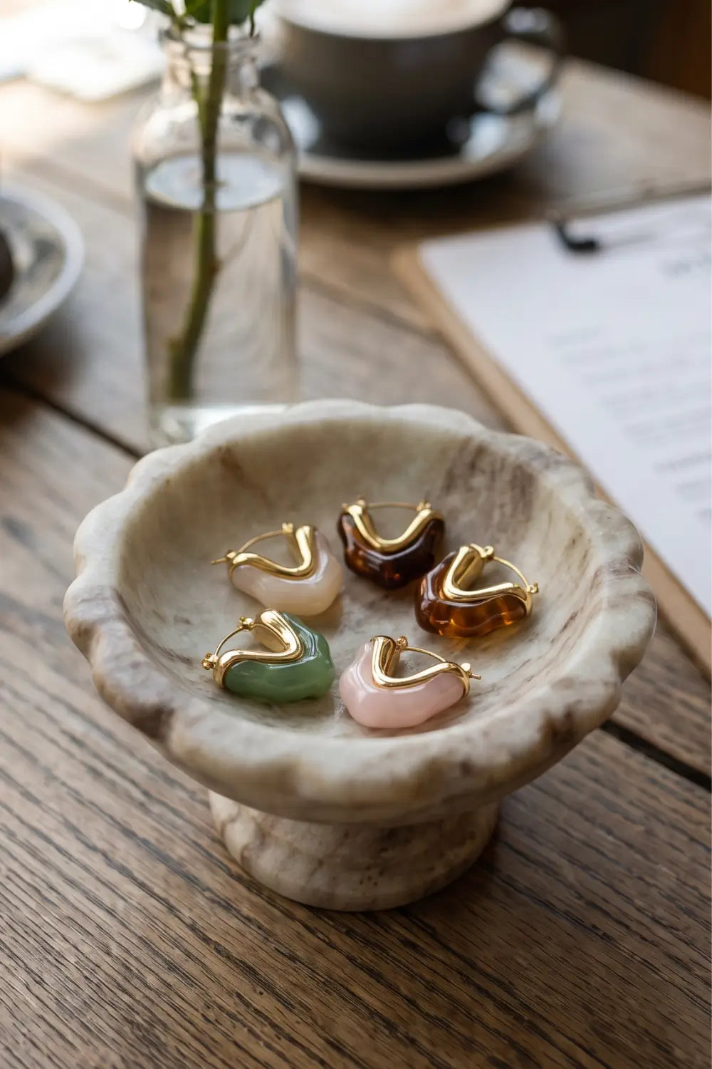 Five small crescent-shaped jewelry pieces in pastel and earthy colors arranged in a shallow stone dish on a wooden table, with a blurred cafe-style background including a cup, glass, and paper.