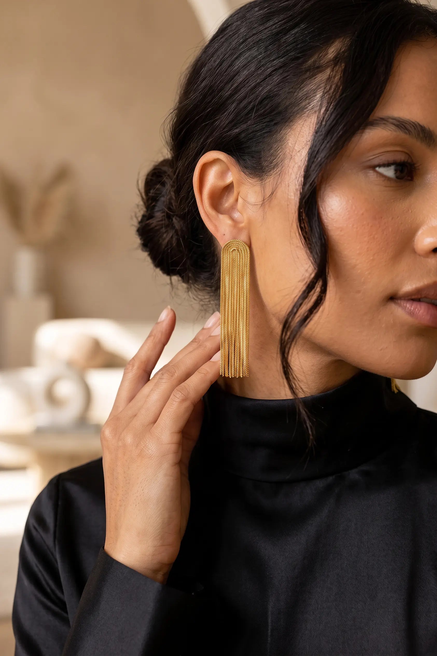 A close-up of a woman wearing a long, gold-toned fringe earring and a black turtleneck top, with a softly blurred warm beige interior in the background.