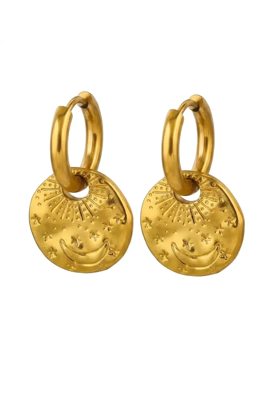 A pair of gold huggie hoop earrings with textured disc charms featuring crescent moon and star designs, shown on a plain white background.