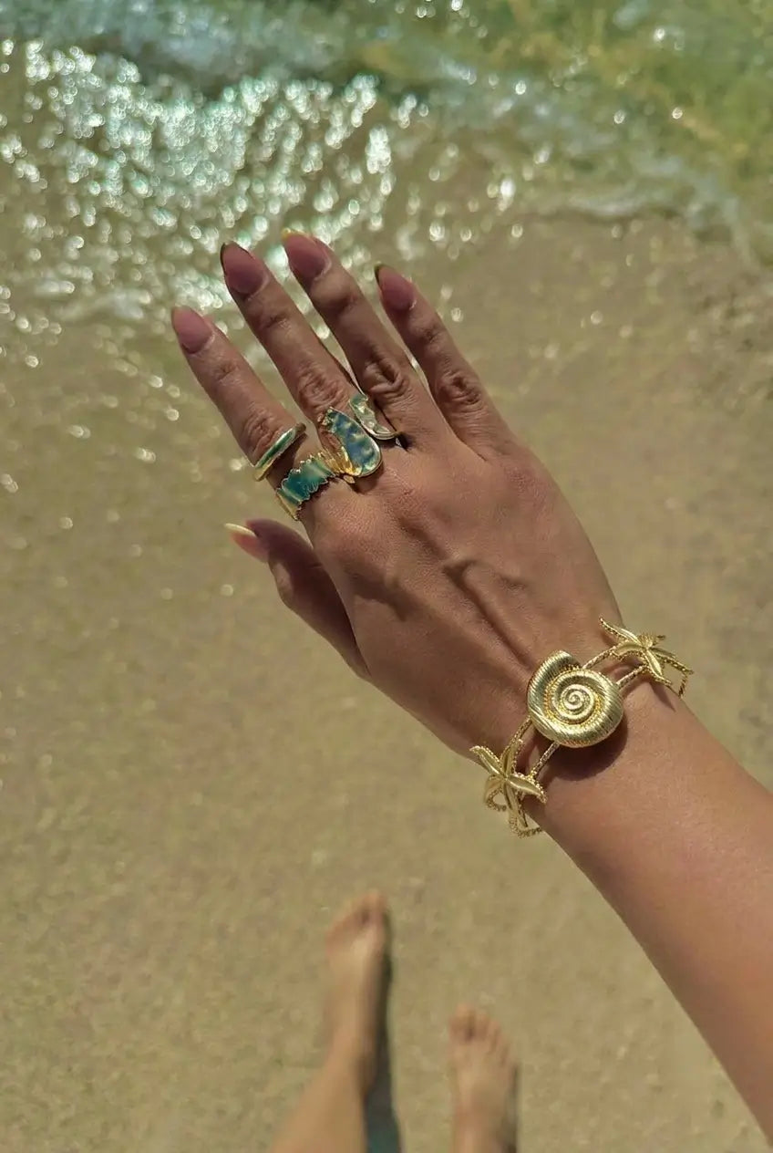 A close-up of a person's hand and wrist at the beach, wearing a chunky gold spiral bracelet and several gold rings with turquoise-colored stones, with wet sand and water in the background.