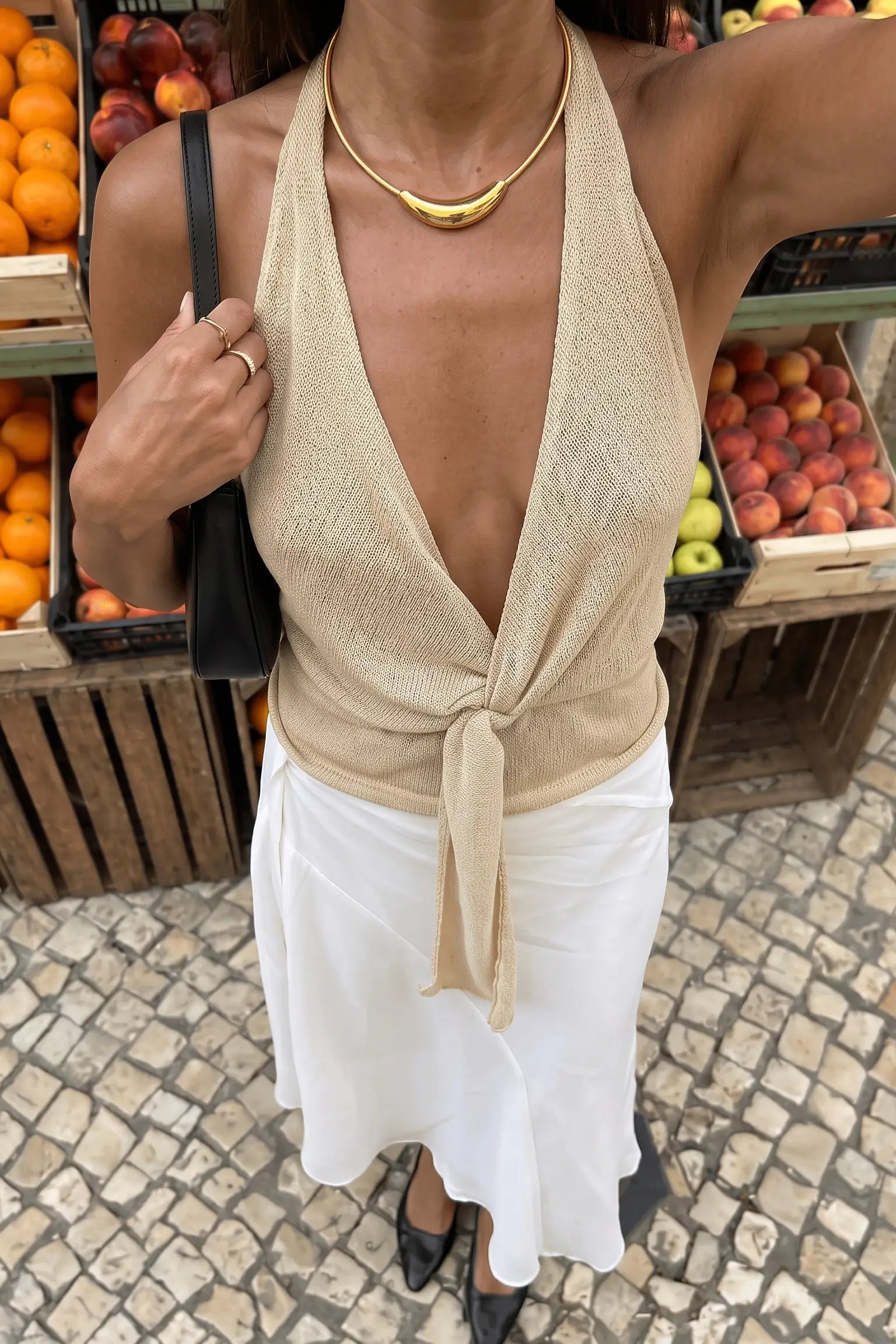 A woman wearing a beige halter-neck knit top with a deep plunging V-neck and front knot detail, paired with a white skirt, standing in front of a fruit stand.