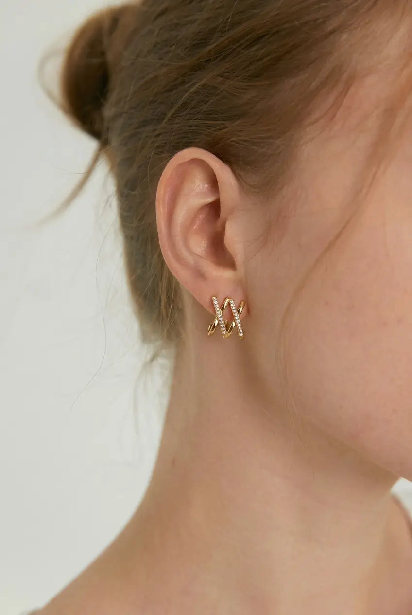 A close-up side profile of a woman's ear wearing a pair of gold hoop earrings with small clear stones, against a plain light background.