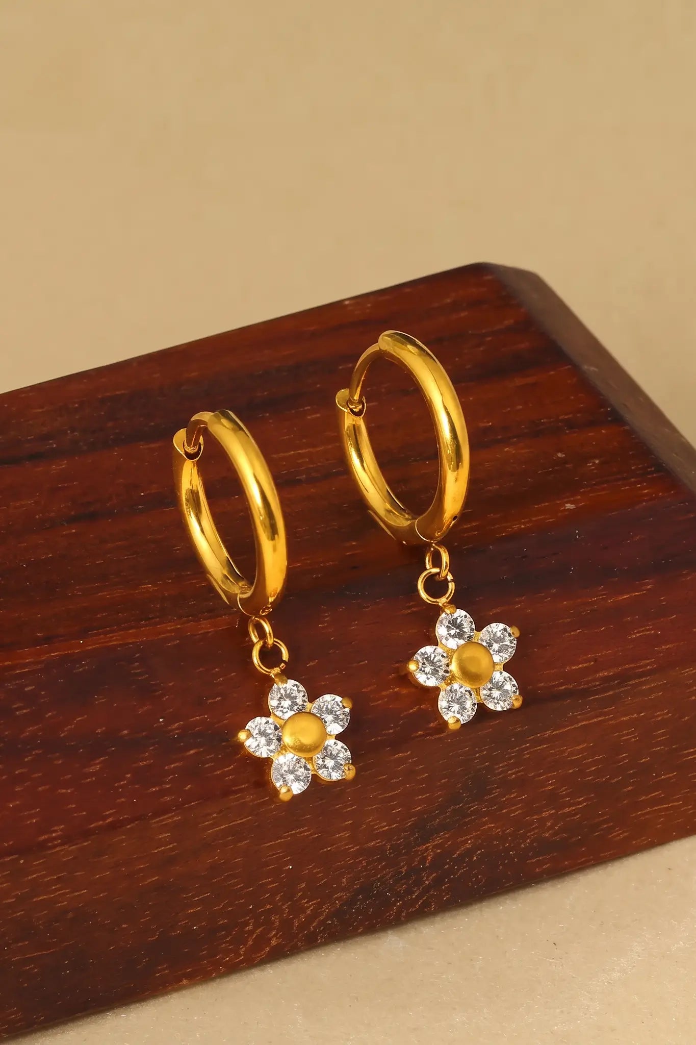 A pair of gold huggie hoop earrings with dangling flower charms, displayed on a dark wooden block against a beige background.