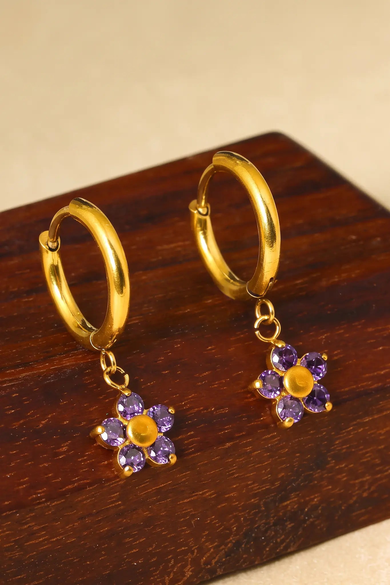 A pair of gold huggie hoop earrings with dangling flower charms made of purple stones and gold centers, displayed on a dark wooden block.