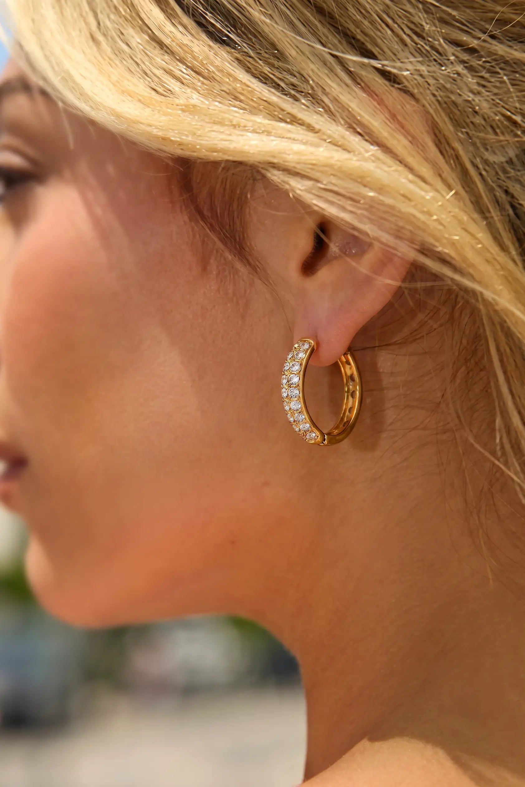 A close-up side profile of a woman wearing a gold hoop earring adorned with small clear stones, set against a softly blurred outdoor background.
