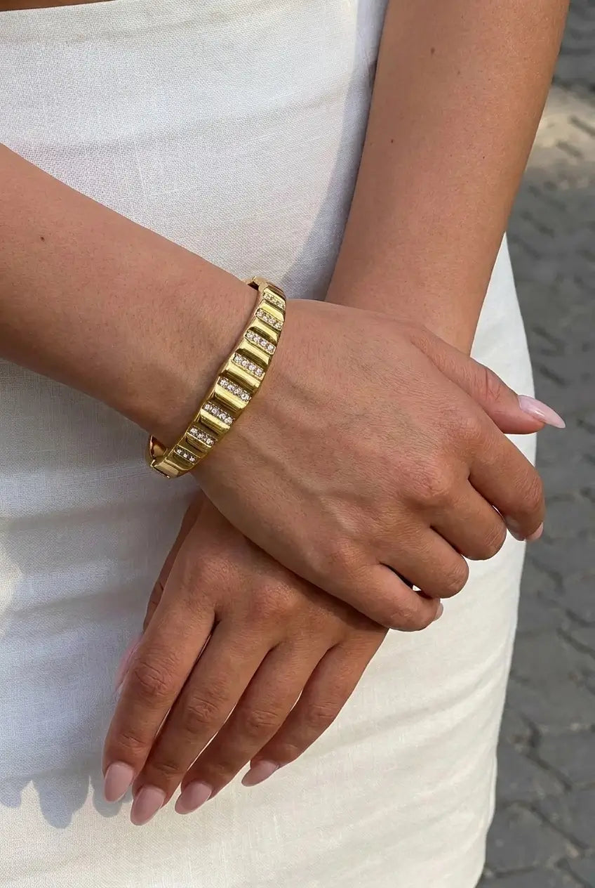 A close-up of a person's hands and arms wearing a gold-toned segmented bracelet with clear stones, set against a white dress and a gray paved background.