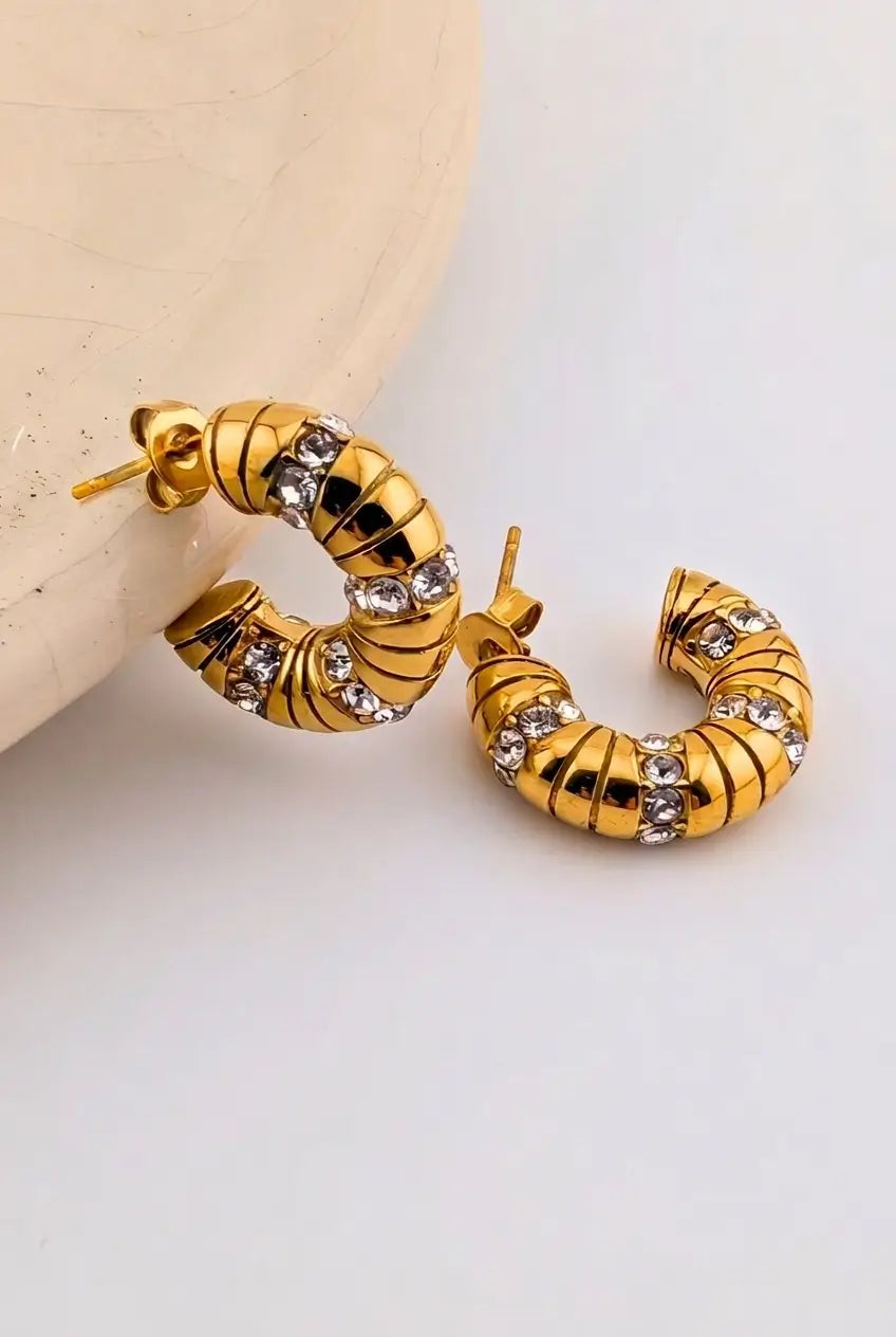 A pair of gold hoop earrings with a ribbed, textured design and clear rhinestone accents, displayed on a light beige surface against a white background.
