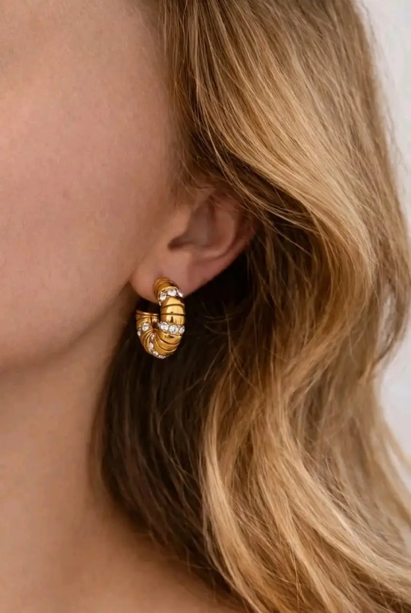 A close-up of a model's ear wearing a gold huggie hoop earring adorned with small clear crystals, set against her blonde hair.