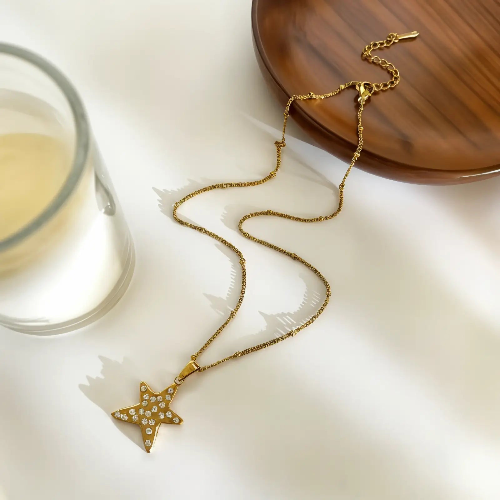 A gold-toned star pendant necklace with small clear stones, displayed on a white surface next to a glass and a wooden plate.