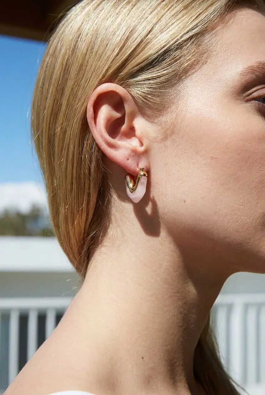 A close-up side profile of a woman wearing a small gold hoop earring with a white oval inlay, photographed outdoors with a blurred background.