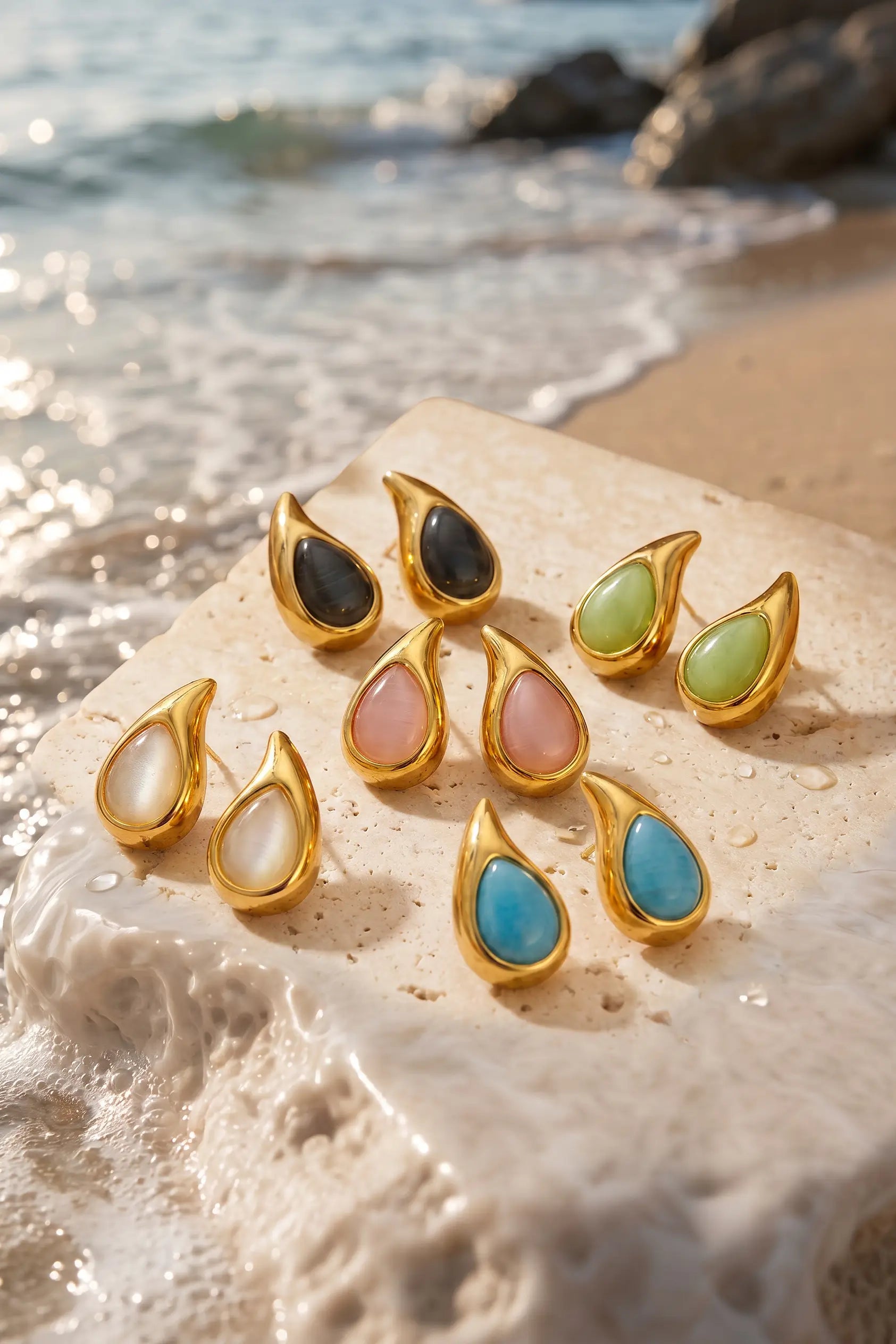 A set of gold teardrop-shaped earrings with colorful stone inlays, arranged on a wet sandy beach with ocean waves in the background. The earrings feature black, light green, pink, white, and blue stones.