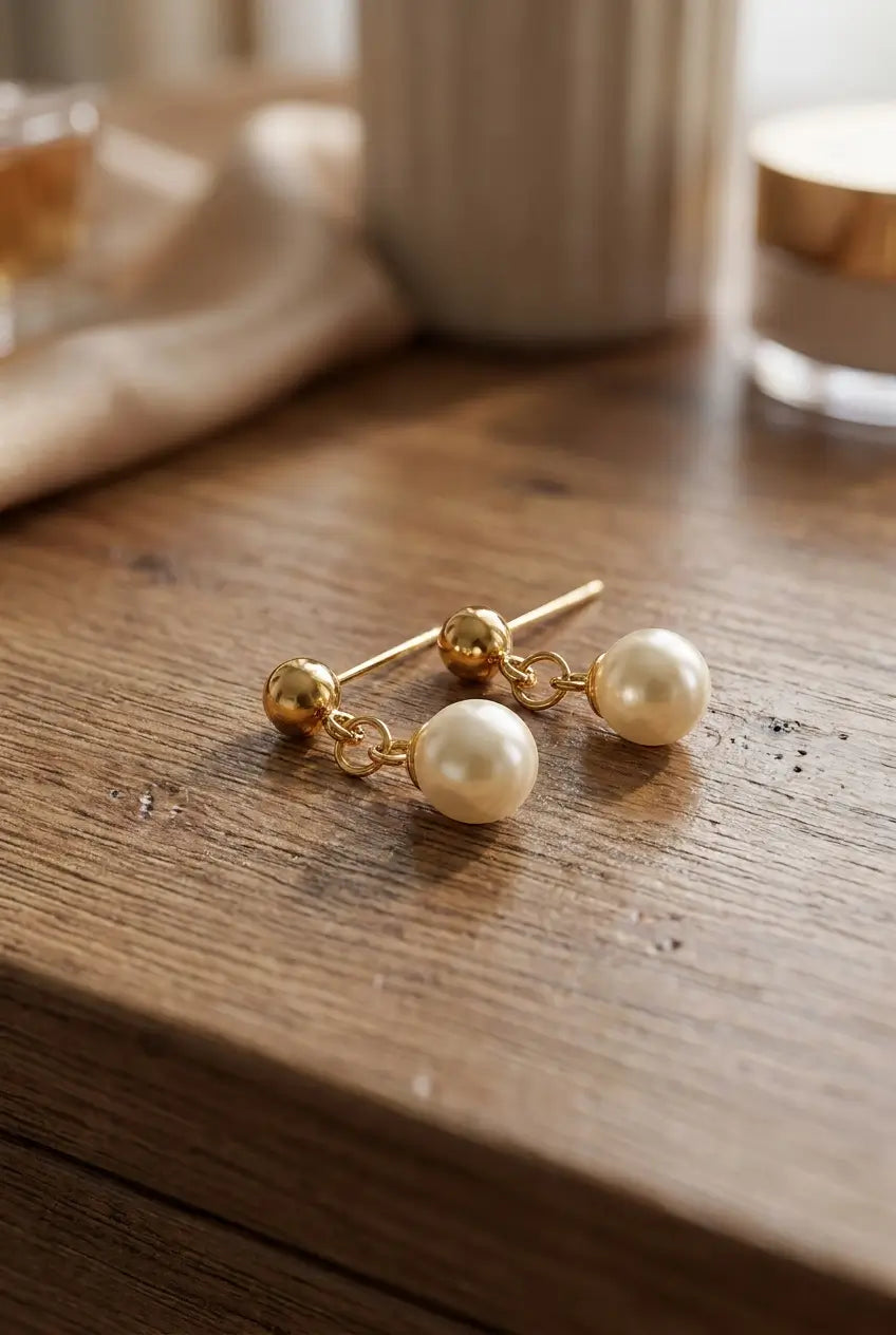 A pair of gold post earrings with round white pearl drops, resting on a rustic wooden tabletop.