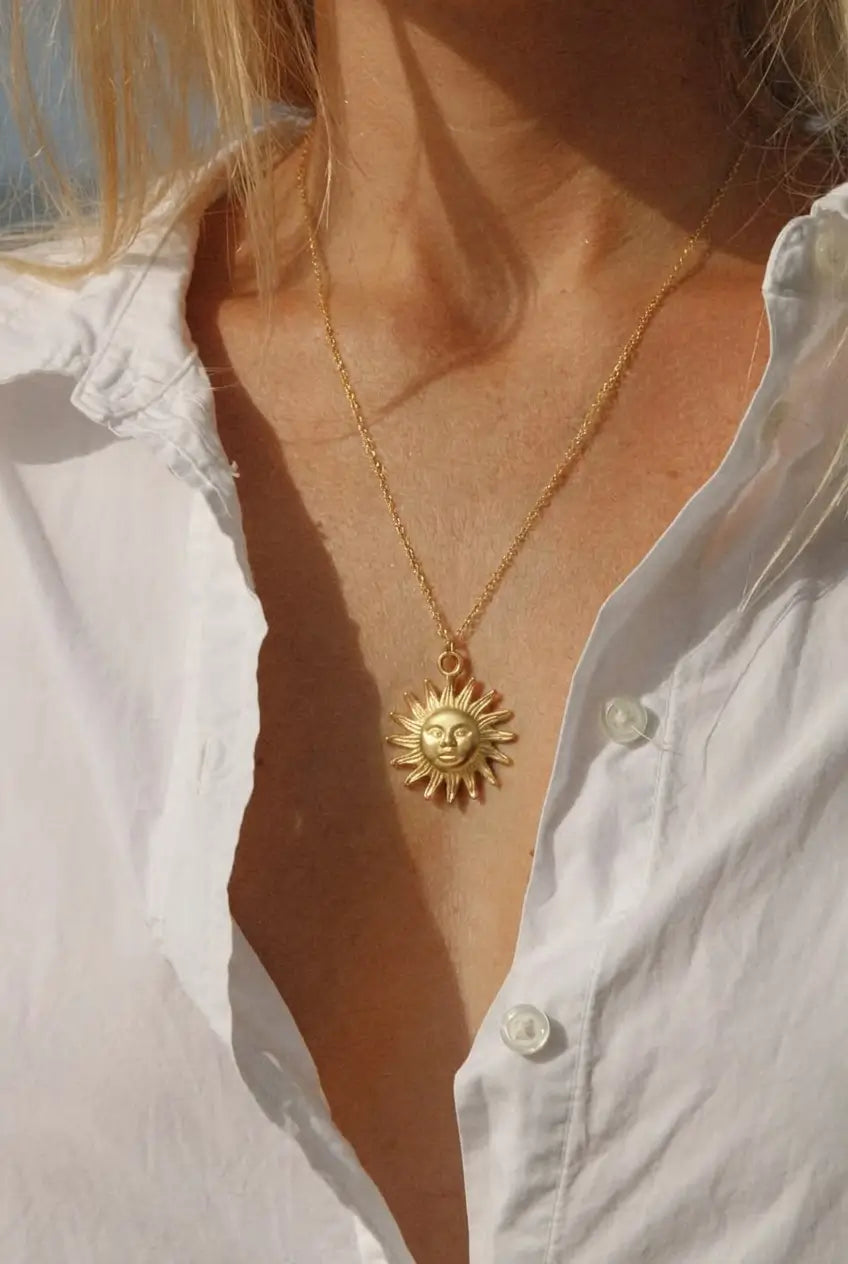 A close-up of a person wearing a delicate gold chain necklace with a sun pendant, styled with an open white button-down shirt.