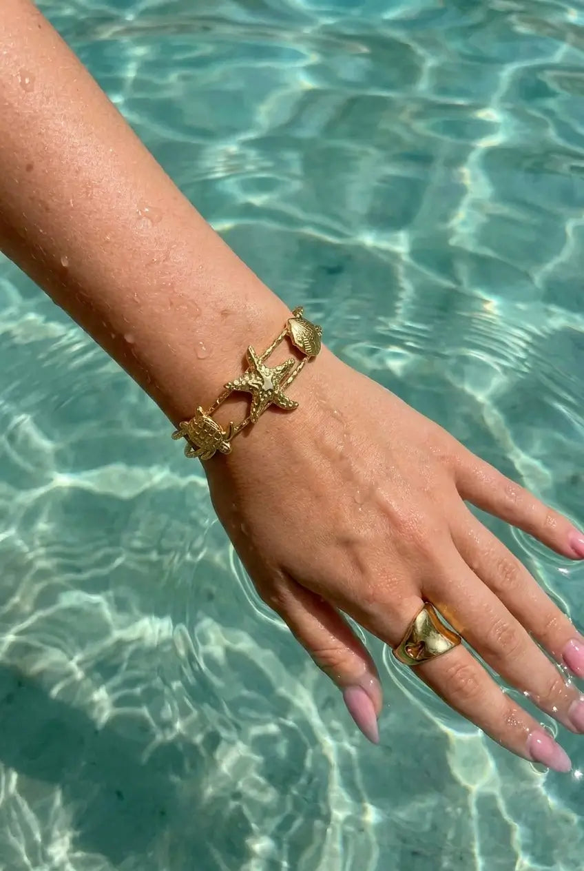 A gold bracelet and matching ring worn on a person's hand, photographed in clear turquoise pool water with sunlight and ripples.