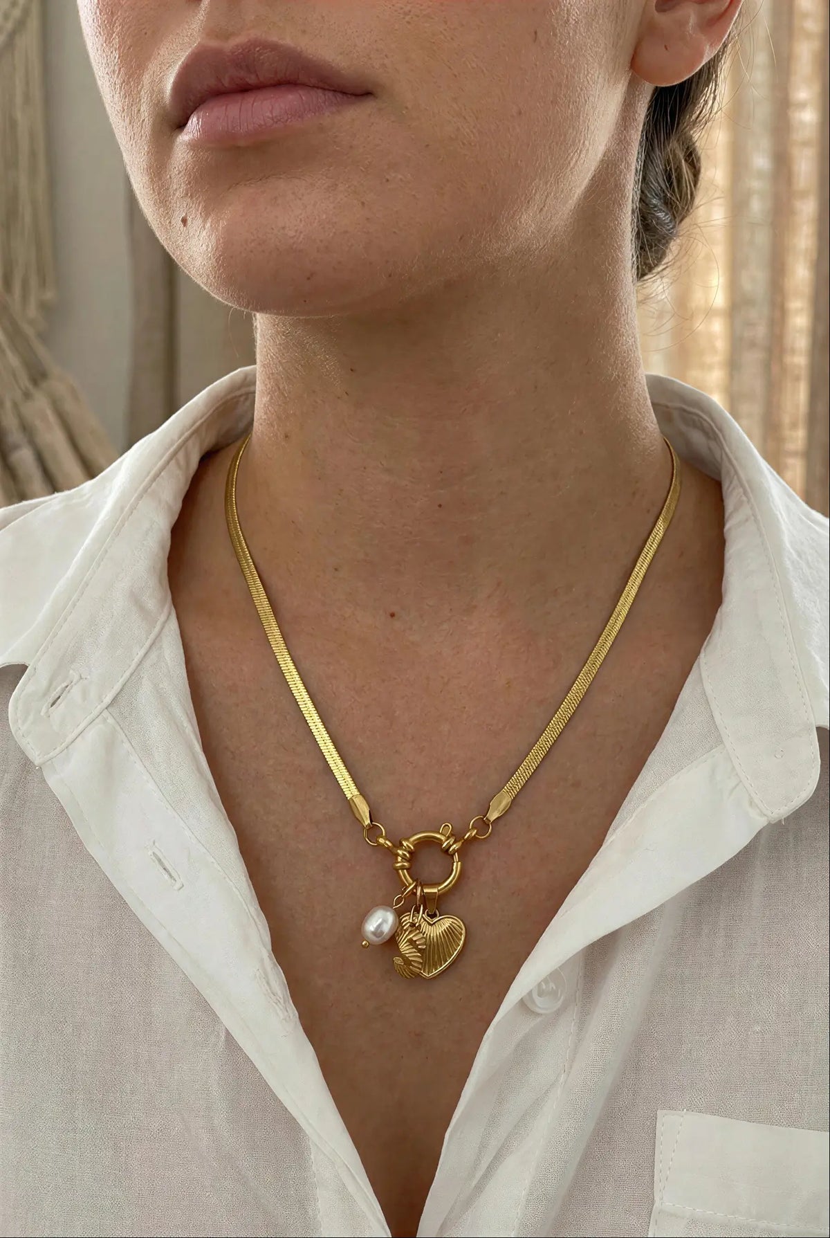A close-up of a model wearing a gold chain necklace with a heart-shaped charm and a small pearl bead, styled with a white collared shirt.