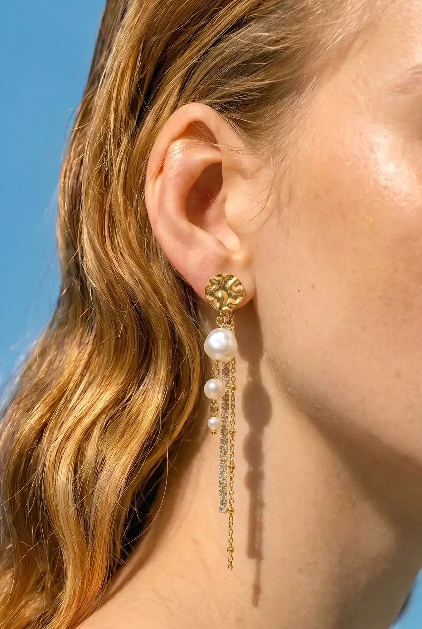 A close-up side profile of a woman wearing a long gold dangle earring with white pearl-like drops and small clear stones, against a bright blue sky.