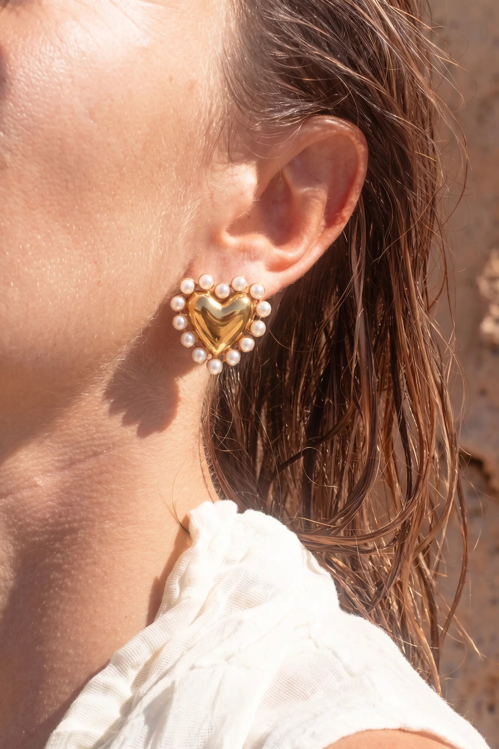 A close-up of a woman's ear wearing a gold heart-shaped stud earring surrounded by small white pearls, with wet brown hair and a white top visible against a warm outdoor background.