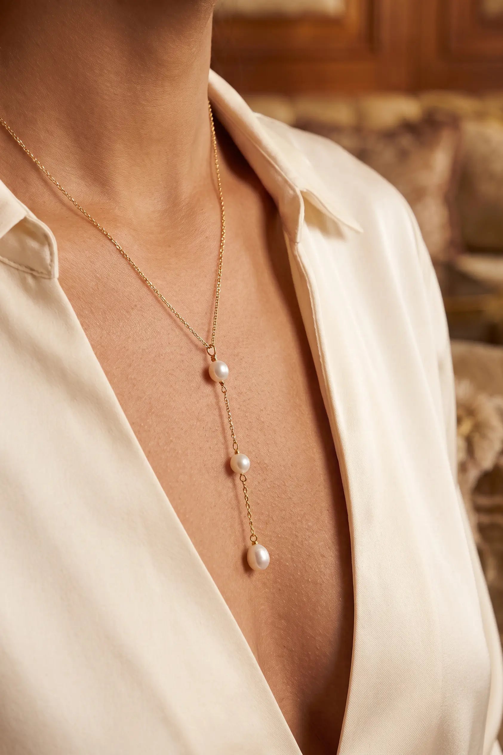 A close-up of a model wearing a delicate gold lariat-style necklace with three white pearl drops, styled with a cream satin shirt.