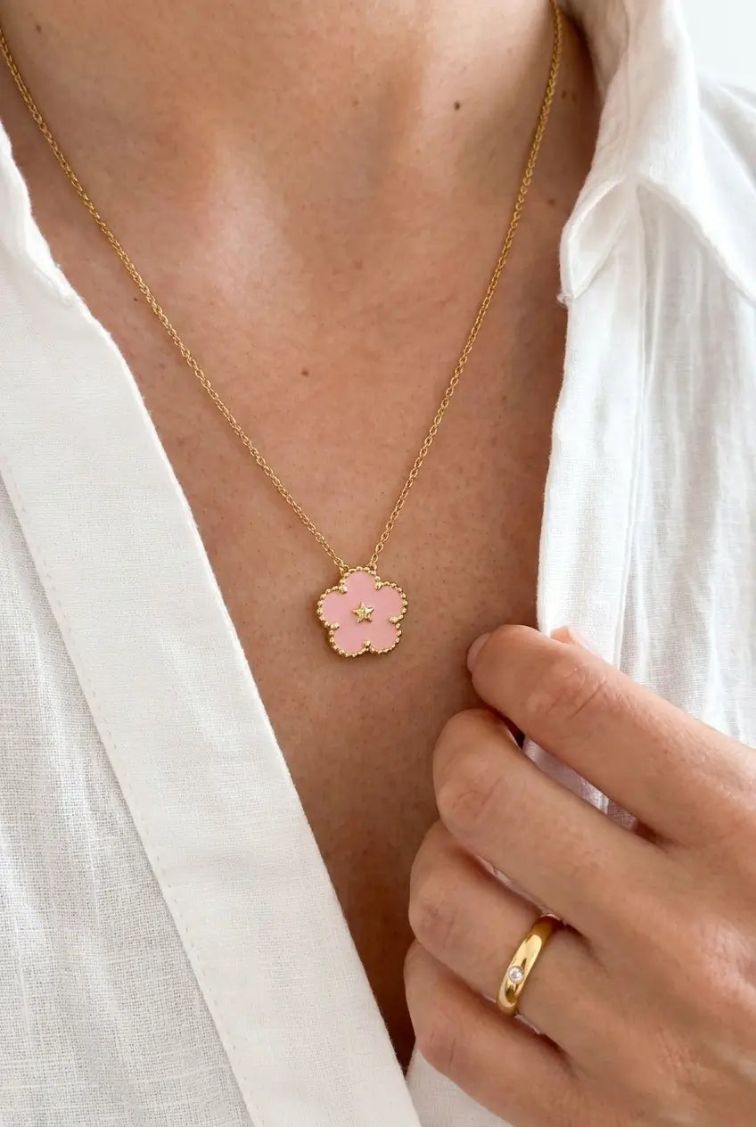 A close-up of a person wearing a delicate gold chain necklace with a small pink flower-shaped pendant, styled with an open white shirt.
