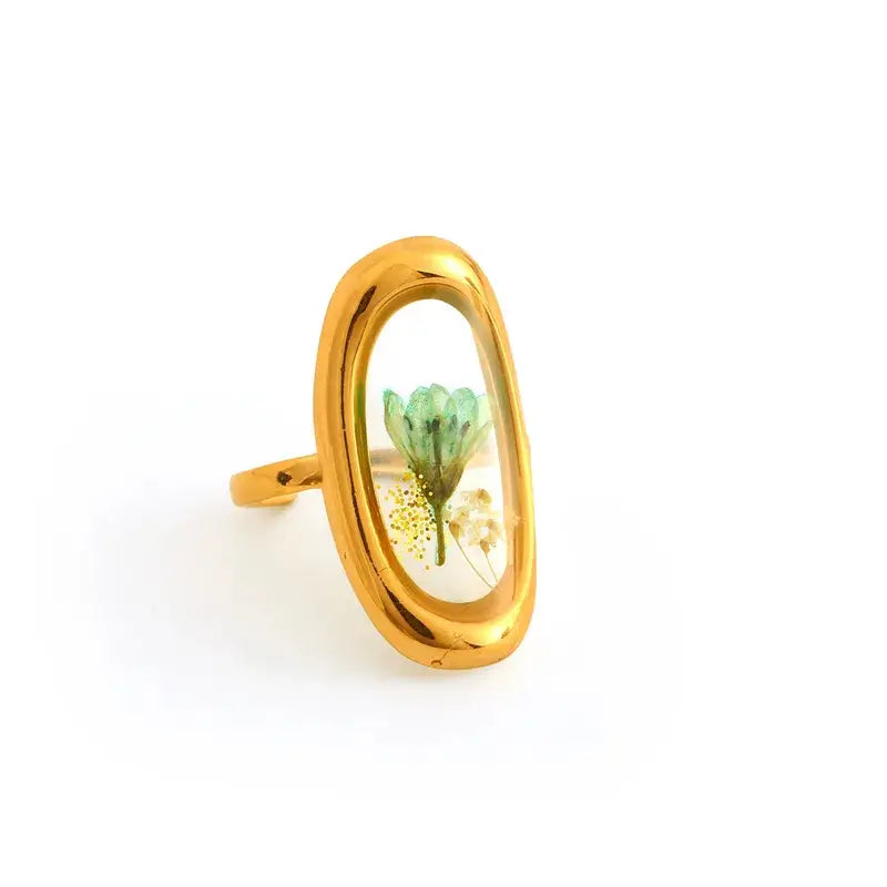 A gold oval ring featuring a clear inlay with a preserved green flower and small white blossoms, shown on a plain white background.