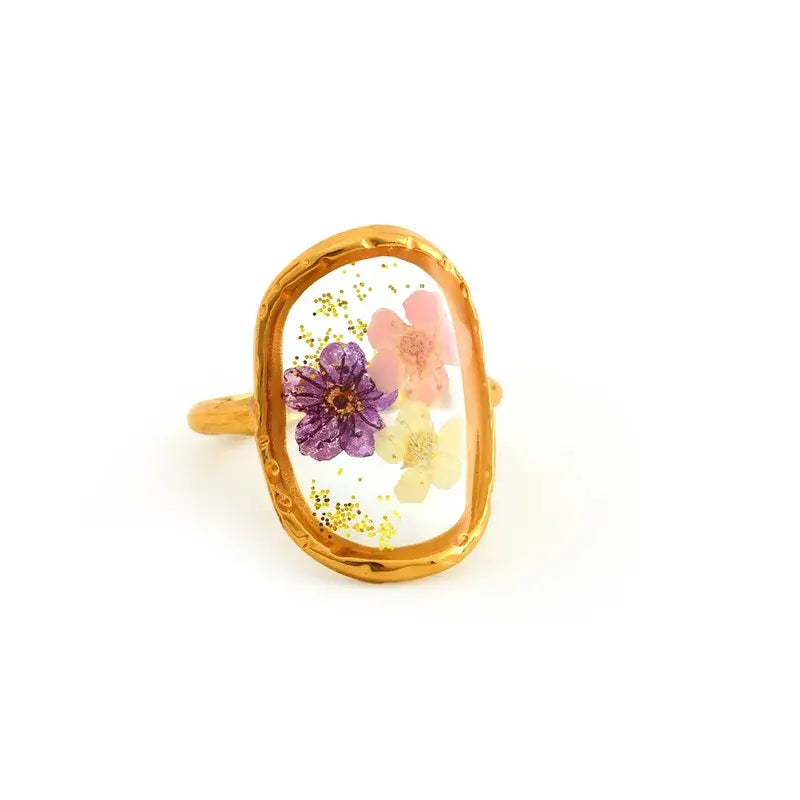 A gold ring with an oval bezel containing a clear resin inlay with dried flowers in purple, pink, and yellow, set against a plain white background.