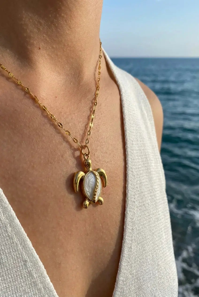 A gold turtle pendant necklace worn by a person outdoors with the ocean in the background. The pendant features a white mother-of-pearl shell center.