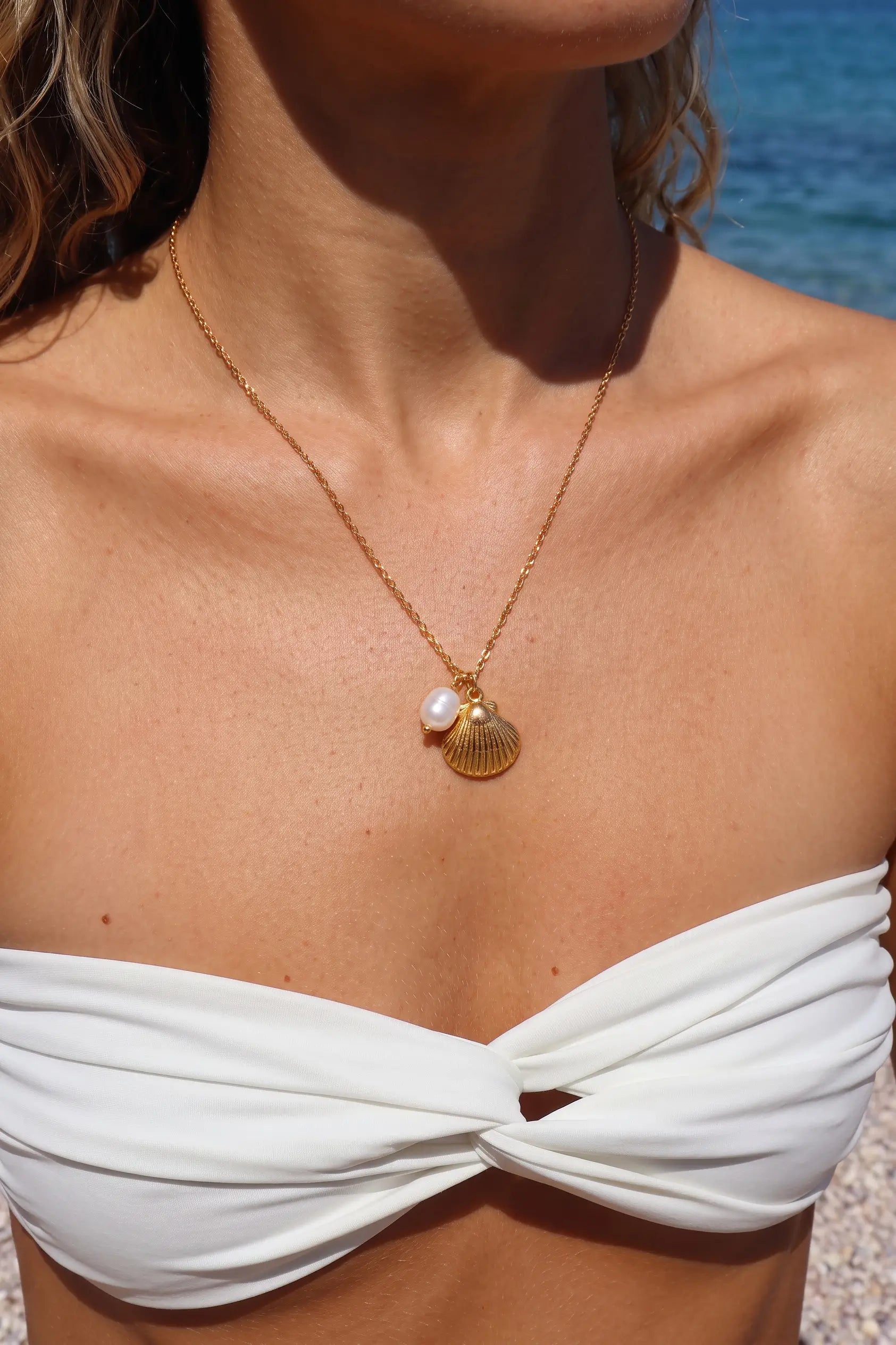 A close-up lifestyle shot of a model wearing a delicate gold chain necklace with a small gold seashell and a white pearl pendant, styled with a white strapless bikini top against a sunny beach background.