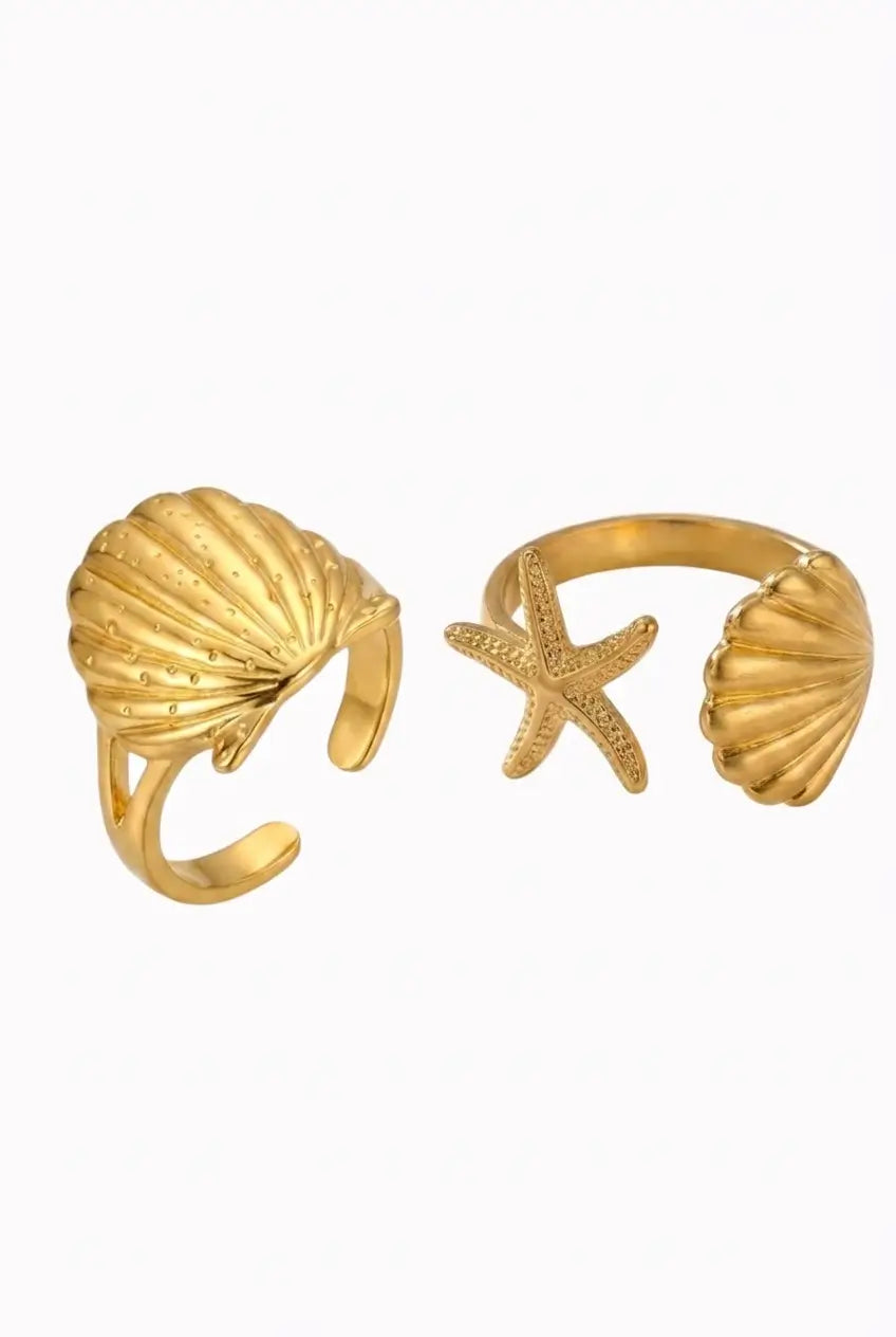 A pair of gold-toned open cuff rings, one featuring a textured seashell and the other a starfish, displayed on a clean white background.