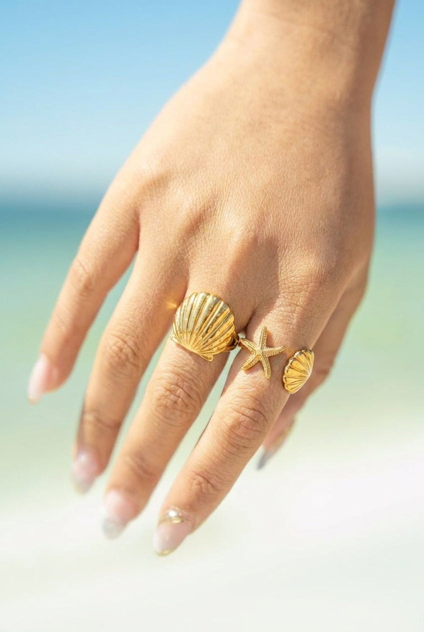 A close-up of a hand wearing three gold-toned seashell and starfish rings against a blurred beach background.