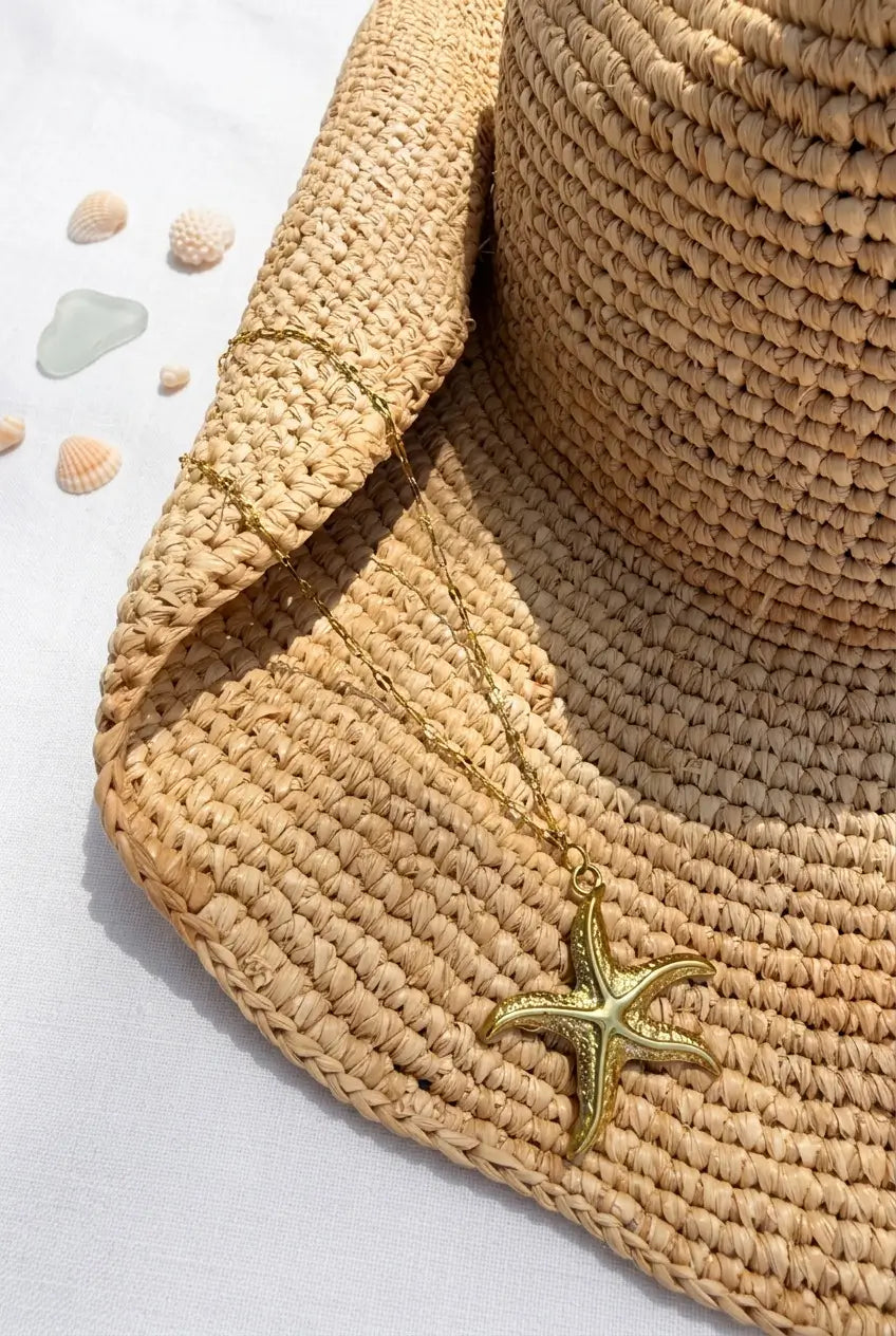 A gold starfish pendant necklace draped over a woven straw hat, with small seashells on a white surface.