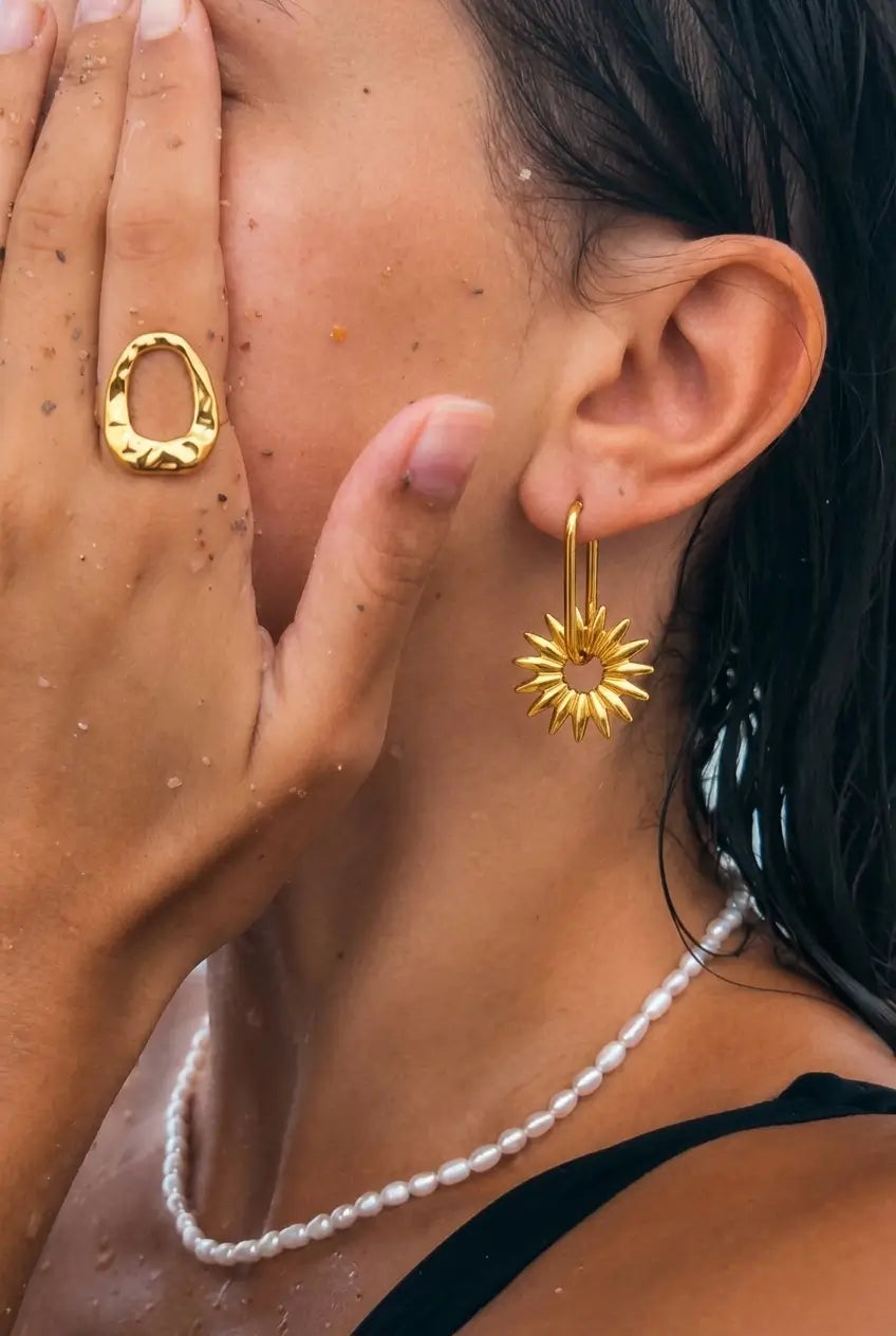 Close-up of a woman wearing a gold sunburst dangle earring, a textured gold ring, and a white pearl necklace.