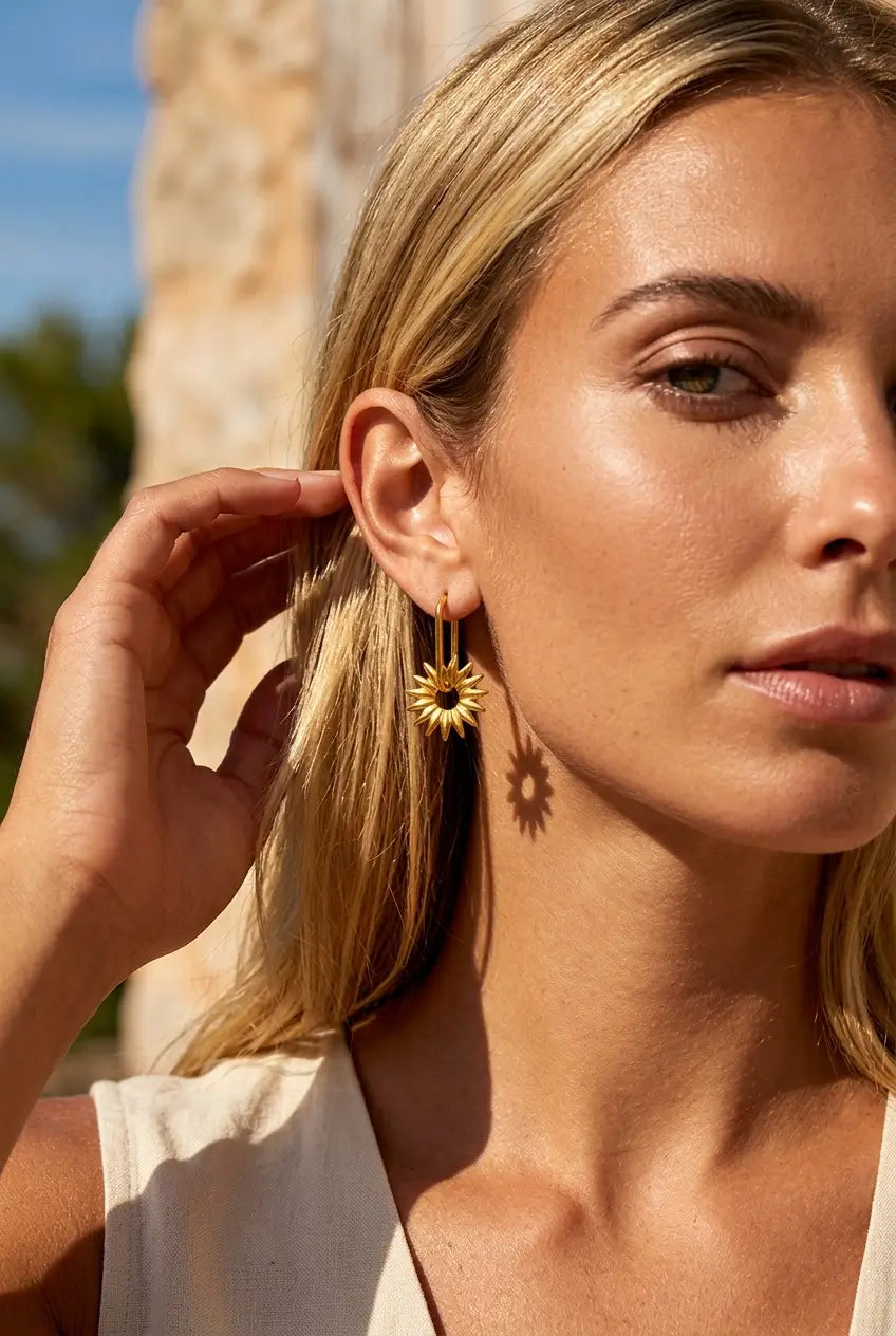A close-up lifestyle shot of a woman wearing a gold sunflower dangle earring outdoors, with warm sunlight and a blurred stone column in the background.