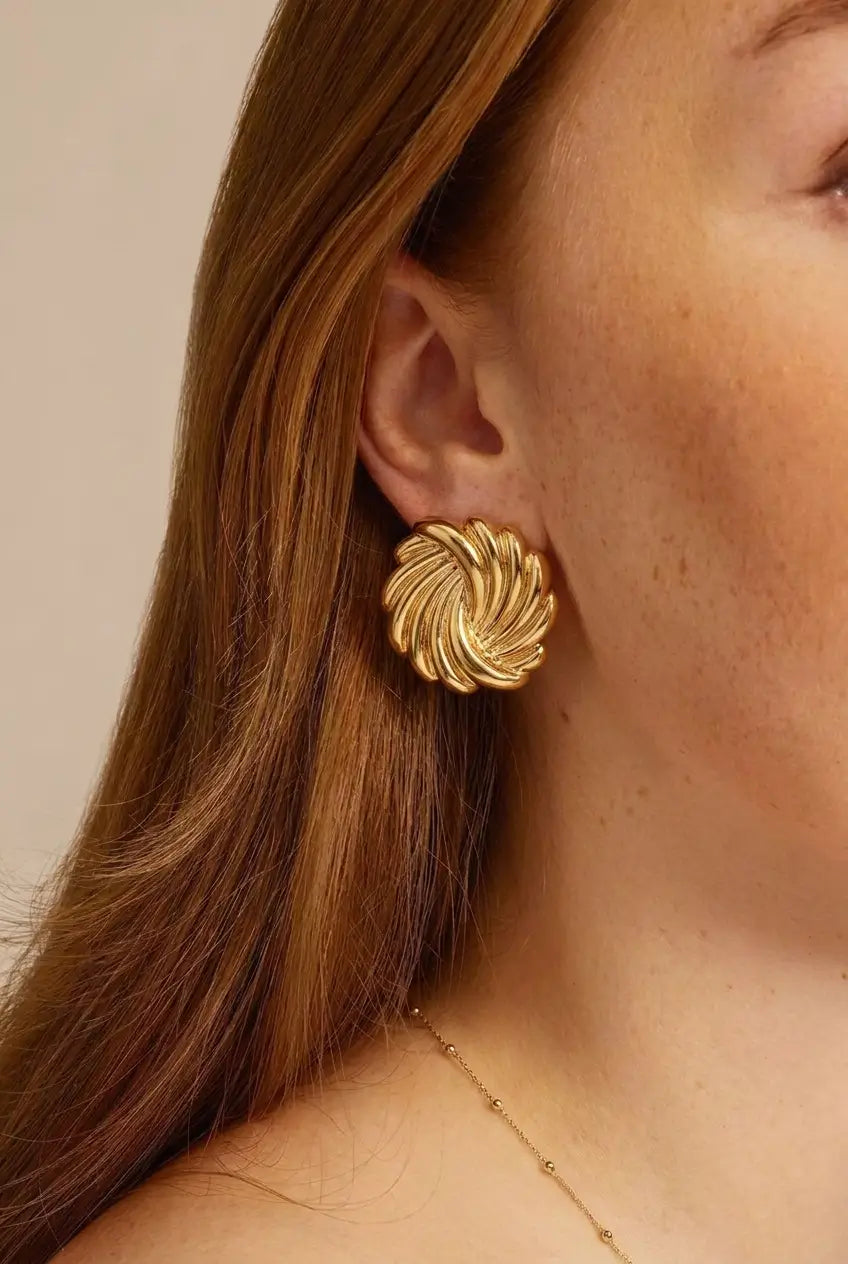 A close-up of a woman's ear wearing a textured, spiral-shaped gold statement earring, with her hair and skin visible against a soft beige background.