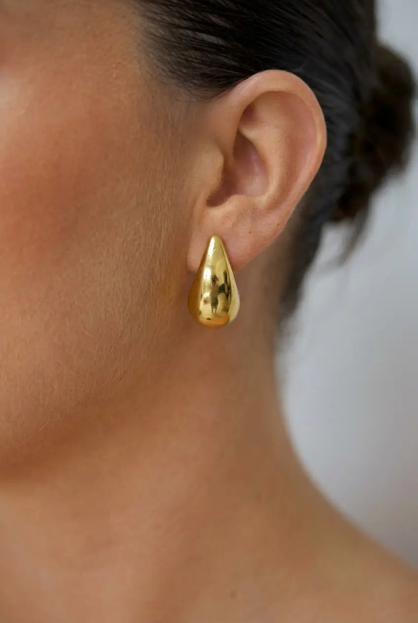 A close-up of a person's ear wearing a small teardrop-shaped gold earring with a textured, hammered finish.