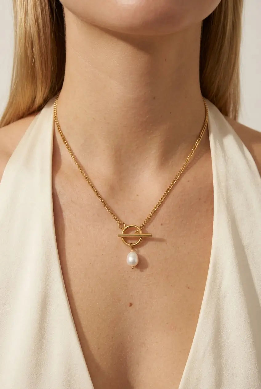 A close-up of a model wearing a gold chain necklace with a toggle ring pendant and a single dangling white pearl, styled with a cream halter top.