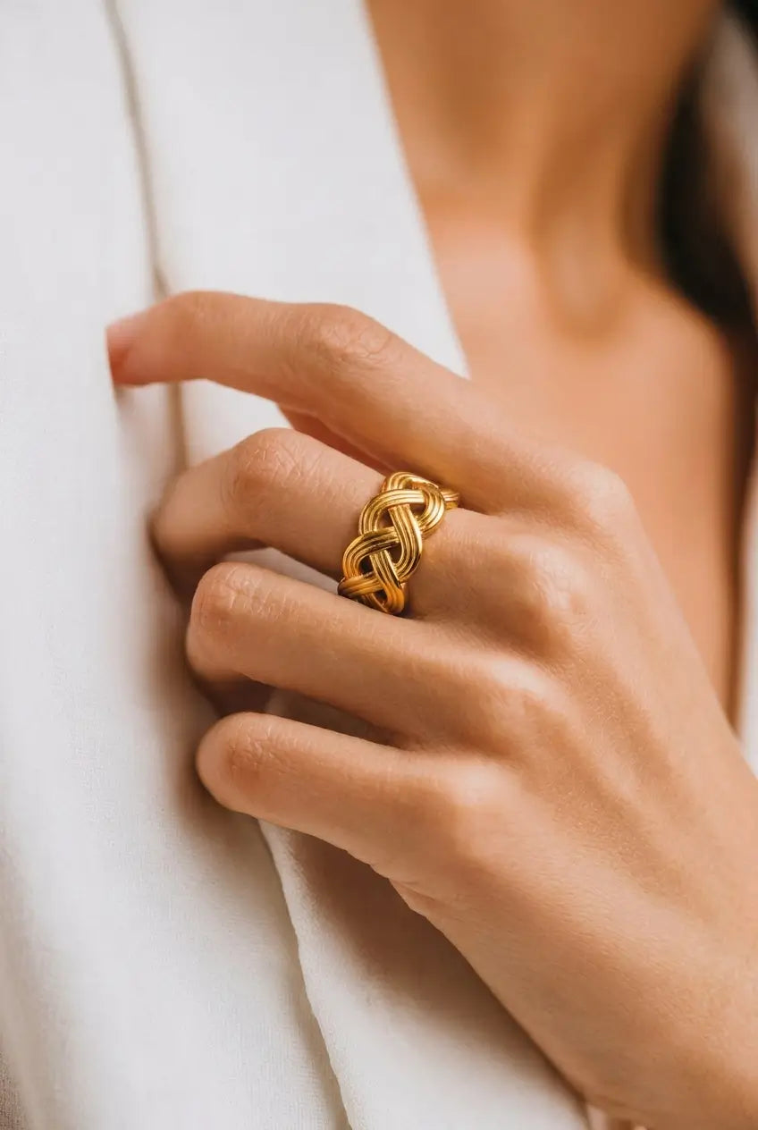A close-up of a person's hand wearing a textured gold ring with an intricate woven knot design, resting against a white garment.