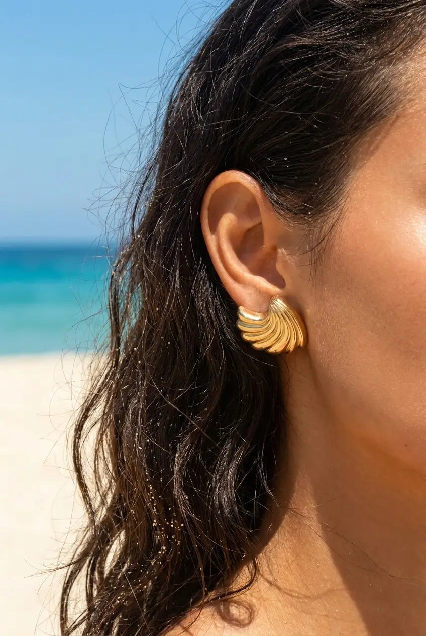 A close-up of a woman's ear wearing a gold-toned, layered scallop-shaped earring, set against a sunny beach background with turquoise water and white sand.