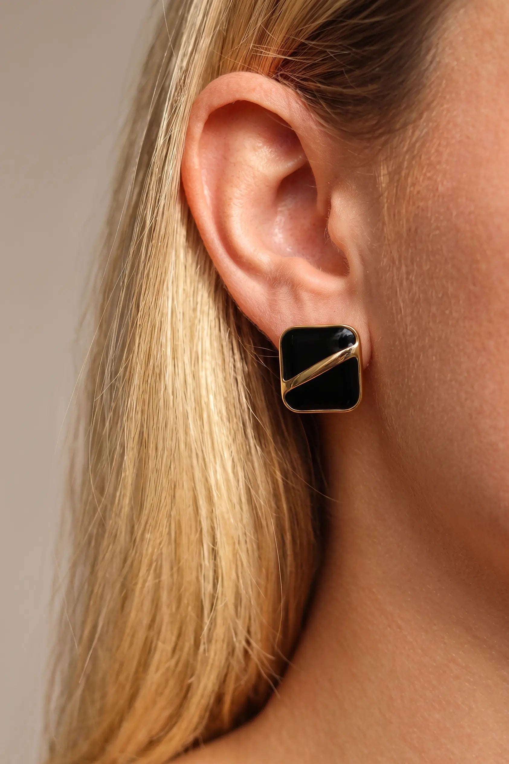 A close-up of a model's ear wearing a square stud earring with a black face and a diagonal gold metal accent.