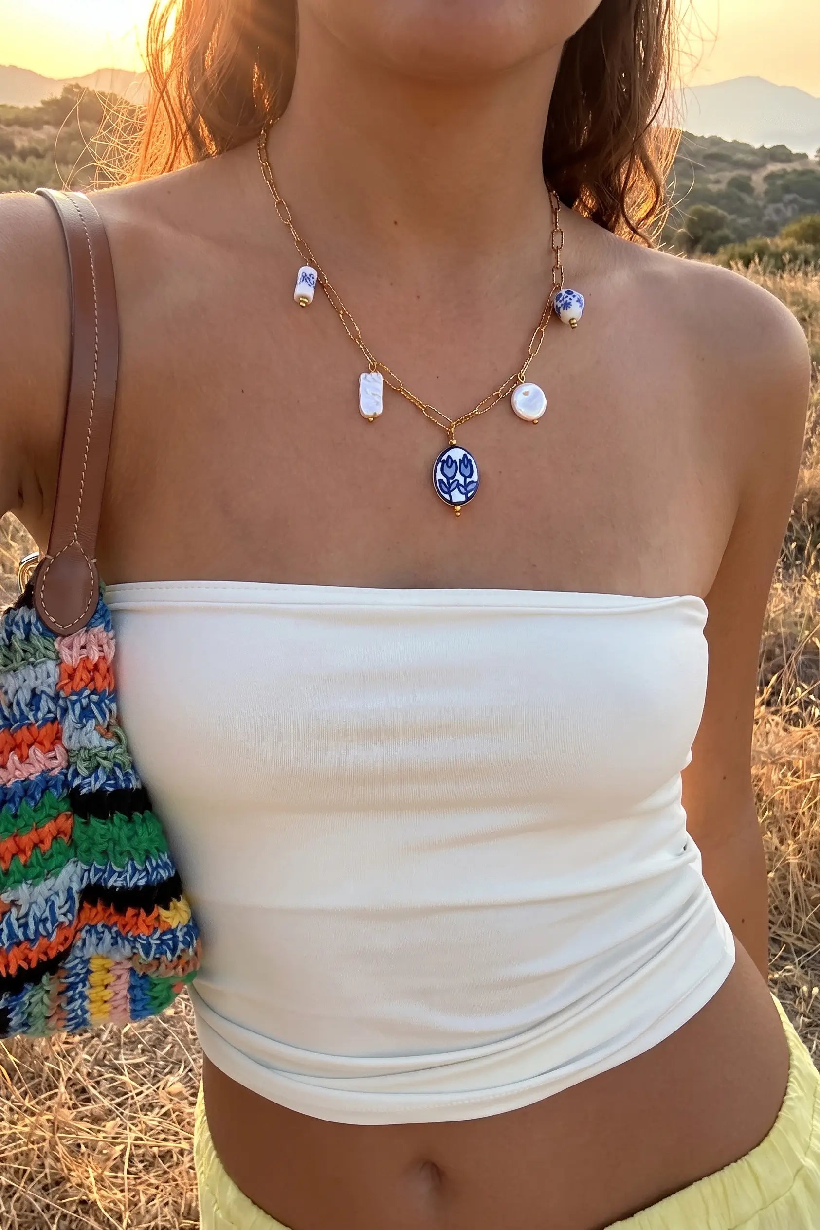 A woman outdoors at sunset wearing a gold-toned necklace with a blue oval pendant and small white and blue charms, paired with a white strapless top and a colorful striped bag.