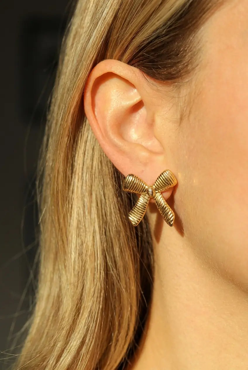 A close-up of a woman's ear wearing a textured gold bow-shaped stud earring, with her blonde hair and skin illuminated by warm sunlight against a dark blurred background.