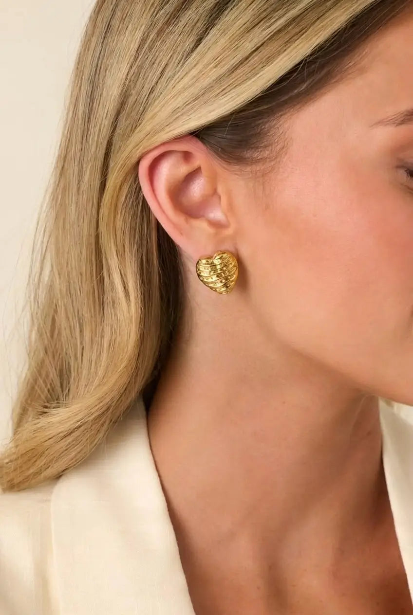 A close-up of a woman's ear wearing a textured, organic-shaped gold stud earring, with her blonde hair and cream-colored top visible against a soft beige background.