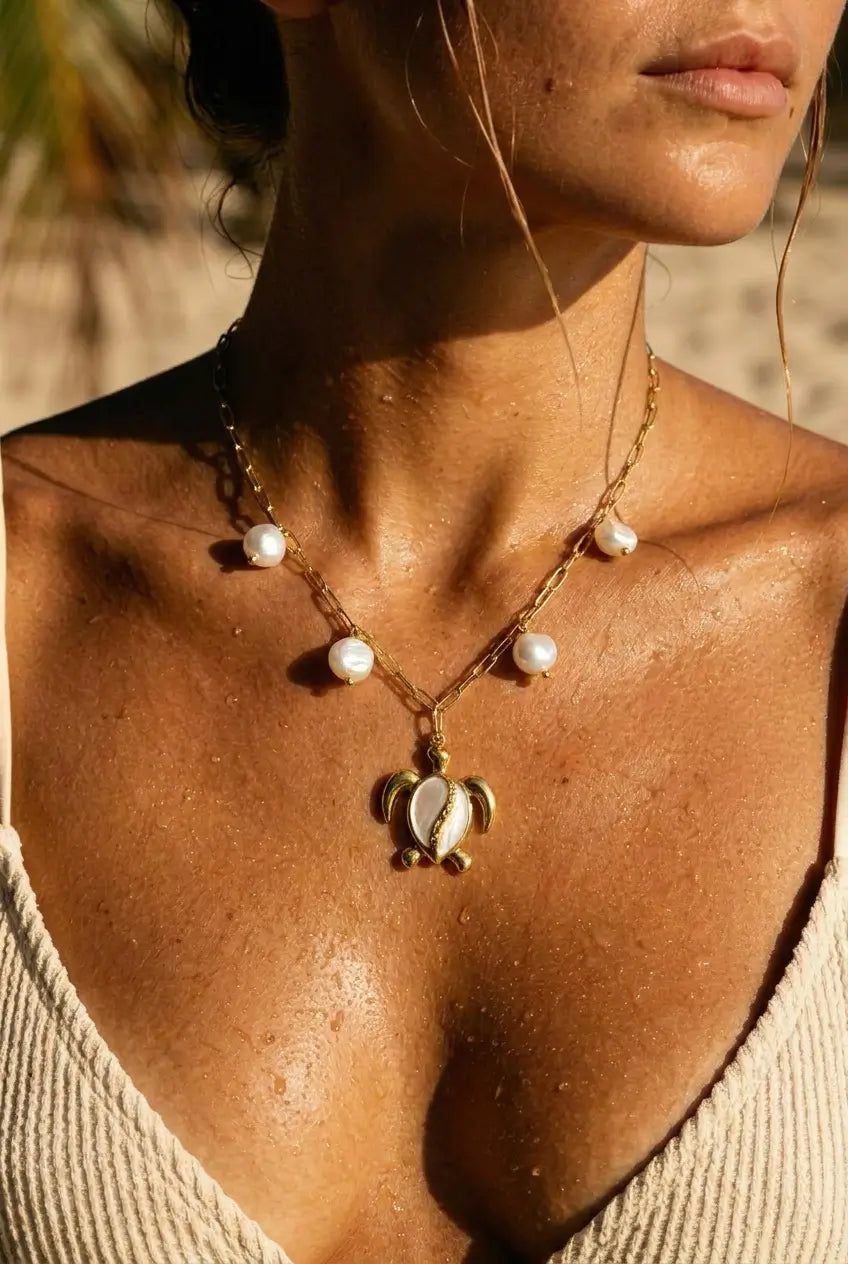 A close-up lifestyle shot of a woman wearing a gold chain necklace with white pearl-like beads and a turtle pendant, set against a warm, sunlit beach background.