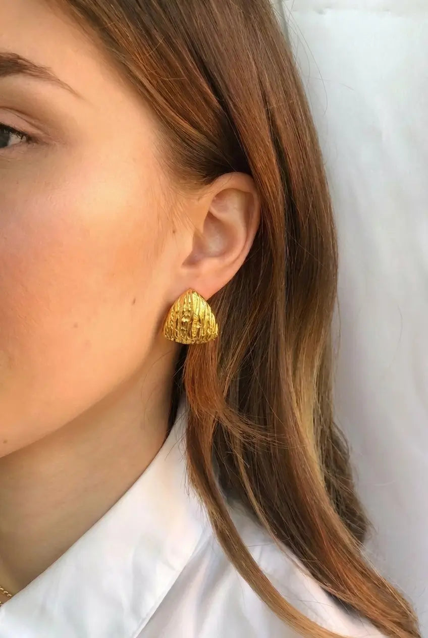 A close-up of a woman's ear wearing a textured, triangular gold stud earring, with her brown hair and white collared shirt visible against a light background.