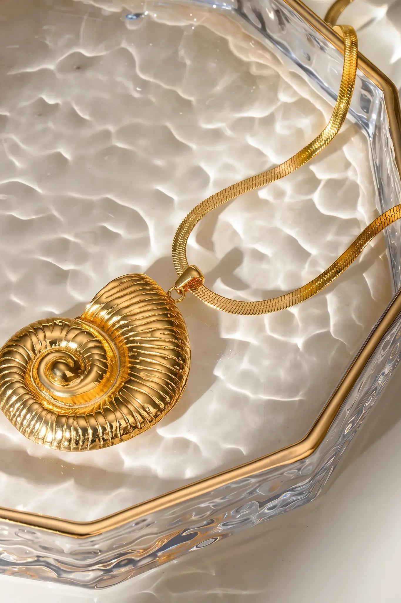 A gold necklace with a textured spiral shell pendant resting on a clear glass dish with a gold-trimmed edge.