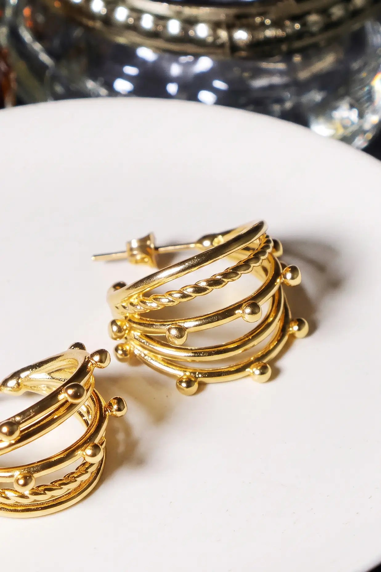 Gold hoop earrings with twisted rope details and small bead accents, displayed on a white surface with a blurred decorative container in the background.
